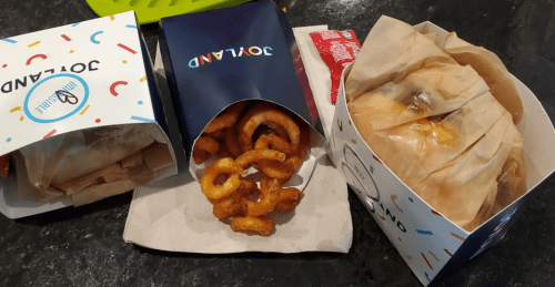 Three takeout containers: one with burgers, one with curly fries, and a packet of ketchup on a dark countertop.