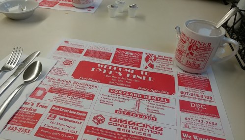 A diner table setting with a menu featuring advertisements and daily specials, alongside a coffee cup and utensils.