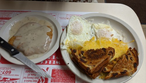 A plate with two fried eggs, hash browns, a biscuit with gravy, and toasted bread on a table with a menu underneath.