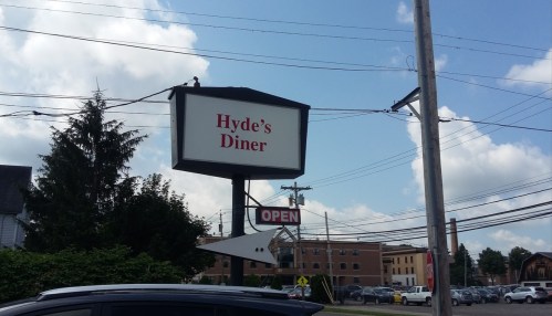 Sign for Hyde's Diner with "OPEN" displayed, surrounded by trees and buildings under a partly cloudy sky.