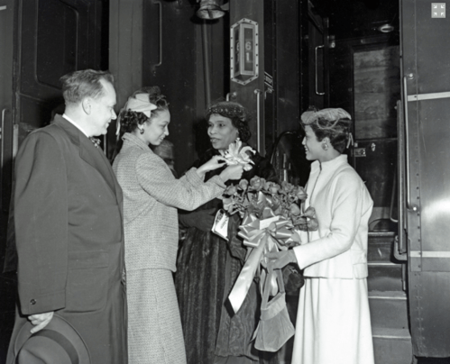 A group of four people, including two women with flowers, greet each other at a train station.