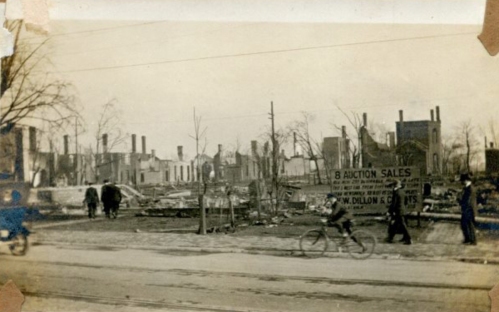 Black-and-white photo of a devastated area with ruins, a sign for auction sales, and people walking and biking.