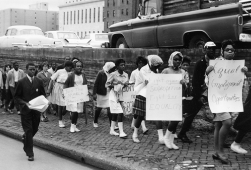 Black-and-white photo of a civil rights march, with participants holding signs for equality and against segregation.