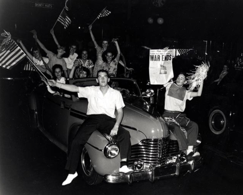 A joyful crowd celebrates in a convertible, waving flags and holding a newspaper announcing the end of war.