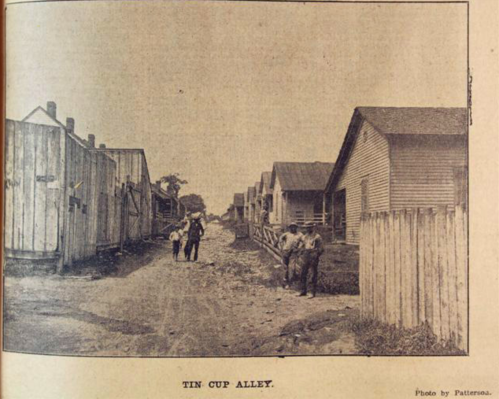 Black and white photo of Tin Cup Alley, featuring wooden houses and people walking along a dirt path.