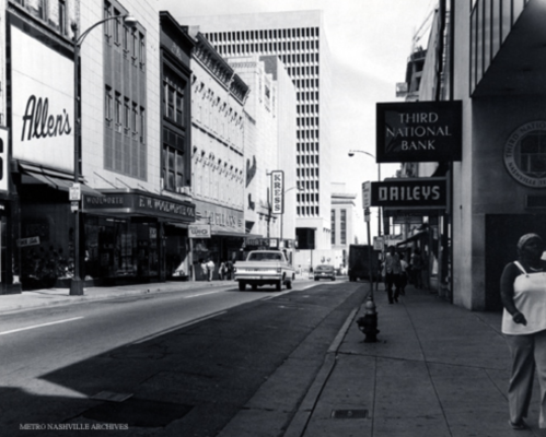 Black and white photo of a city street featuring storefronts, a bank, and a parked car.