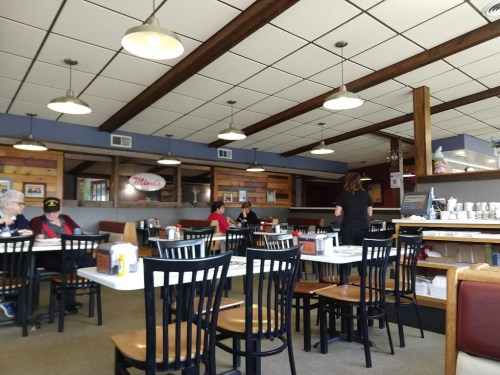 A cozy diner interior with wooden beams, tables, and patrons enjoying their meals. A waitress serves in the background.