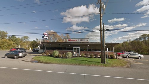 A drive-in restaurant with a sign reading "Mimi's Drive-In," surrounded by cars and trees under a blue sky.