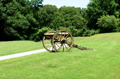 A historic cannon on a grassy field, surrounded by trees and a clear blue sky.