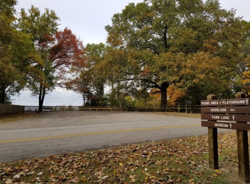 A scenic park area with autumn trees, a sign for picnic and playground, and a view of a lake in the distance.