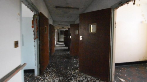 A dimly lit hallway with open doors, scattered debris on the floor, and peeling paint on the walls.