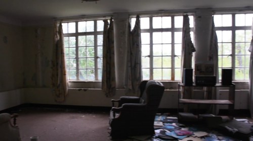 Abandoned room with large windows, tattered curtains, a chair, and scattered papers on the floor.