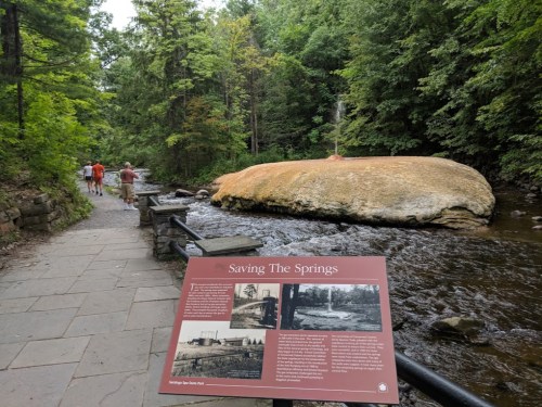 A scenic view of a stream with a large rock, surrounded by trees, and a historical sign about the springs nearby.
