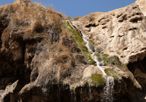 A waterfall cascading down rocky cliffs, surrounded by dry grass and a clear blue sky.