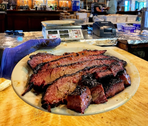 A plate of sliced smoked brisket held by a gloved hand, with a bar and staff in the background.