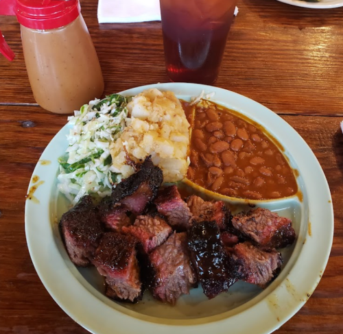 A plate of barbecue beef, coleslaw, potato salad, and baked beans, with a drink and sauce on the side.