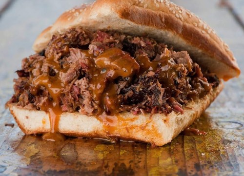 A close-up of a sesame seed bun filled with shredded meat and drizzled with sauce, resting on a wooden surface.