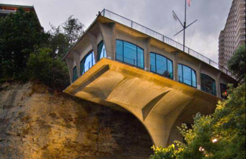 A modern building with large windows, perched on a cliff, supported by a concrete structure, surrounded by greenery.