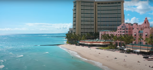A sunny beach scene with turquoise water, palm trees, and a pink hotel along the shoreline.