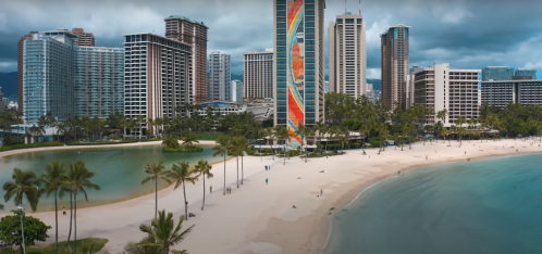 Aerial view of a beach with palm trees, high-rise buildings, and a calm lagoon under a cloudy sky.