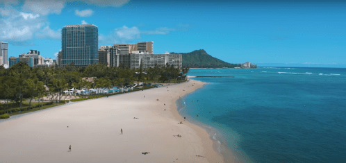A scenic beach with soft sand, clear blue water, and tall buildings in the background, featuring a mountain in the distance.