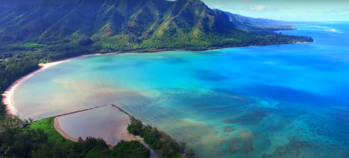 Aerial view of a vibrant blue ocean meeting a lush green coastline and mountains under a clear sky.