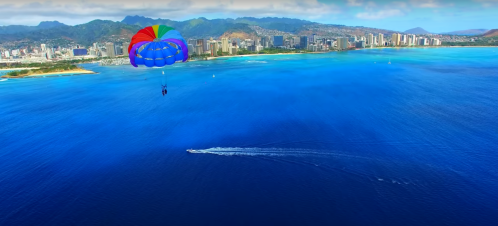 A colorful parachute glides over a vibrant blue ocean near a city skyline and mountains in the background.
