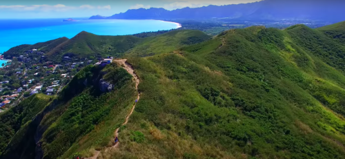 Aerial view of lush green hills leading to a coastline with blue ocean and distant mountains.