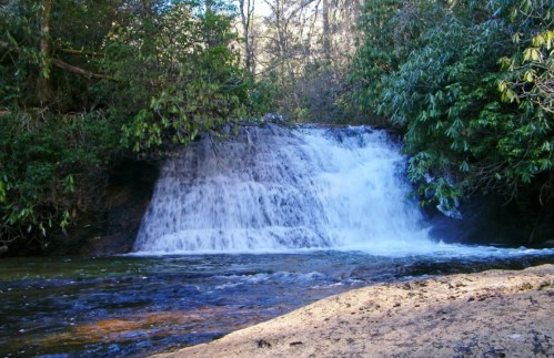 A serene waterfall cascades over rocks into a clear pool, surrounded by lush greenery and trees.