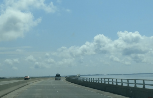 A highway stretches over water under a blue sky with fluffy clouds, with vehicles traveling in the distance.