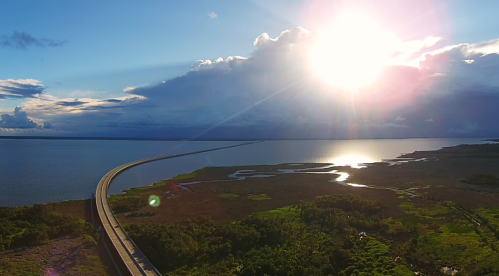 Aerial view of a winding road by a shimmering body of water, with a bright sun and clouds in the sky.