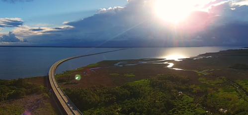 Aerial view of a winding road along a body of water, with sunlight reflecting on the surface and lush greenery nearby.