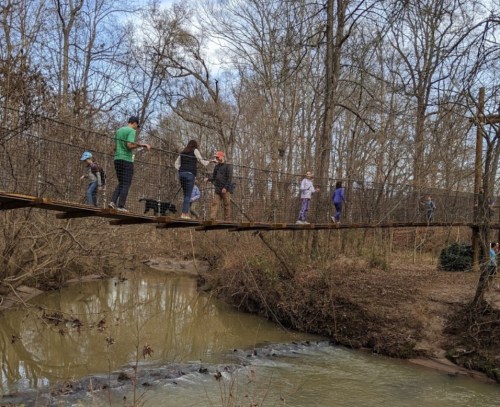 A group of people crossing a suspension bridge over a creek in a wooded area. Trees surround the scene.