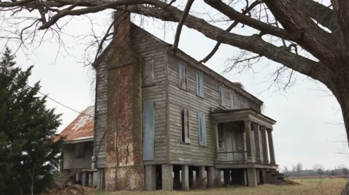 A weathered, two-story wooden house with boarded windows, standing on stilts, surrounded by trees in a rural setting.