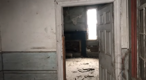 An abandoned room with peeling walls, debris on the floor, and a doorway leading to another dimly lit space.