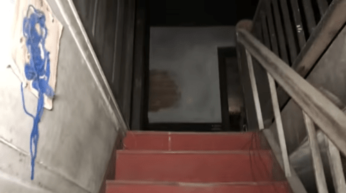 A dimly lit staircase with red steps leading to a dark doorway at the top, with a blue paint splash on the wall.
