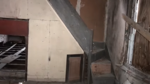 An interior view of a dilapidated space featuring a staircase and exposed walls, with debris on the floor.