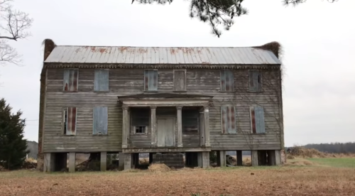 Abandoned two-story wooden house with a rusty roof, boarded windows, and overgrown vegetation in a rural setting.