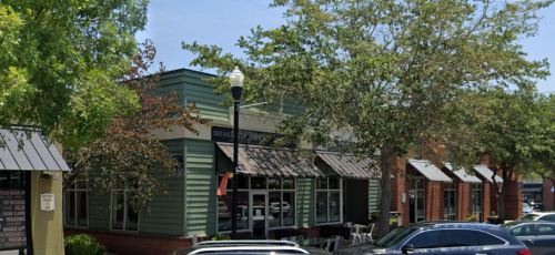 A street view of a green building with awnings, surrounded by trees and parked cars on a sunny day.