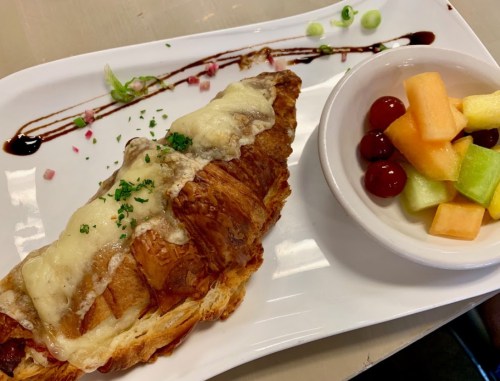 A croissant topped with melted cheese, drizzled with sauce, served with a side of mixed fruit and garnished with herbs.