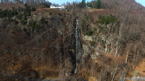 Aerial view of a waterfall cascading down a rocky cliff, surrounded by bare trees and a house on the hillside.