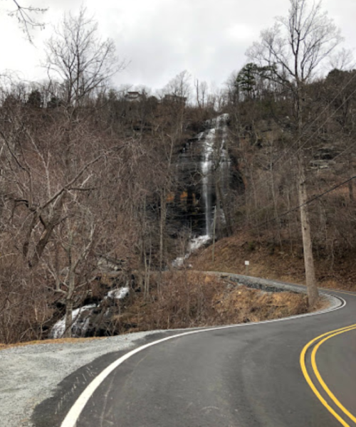 A winding road beside a waterfall cascading down a rocky cliff, surrounded by bare trees and overcast skies.