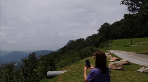 A person stands on a scenic overlook, taking a photo of a mountain landscape under a cloudy sky.