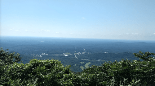 A panoramic view of rolling hills and a distant highway under a clear blue sky, framed by green foliage.
