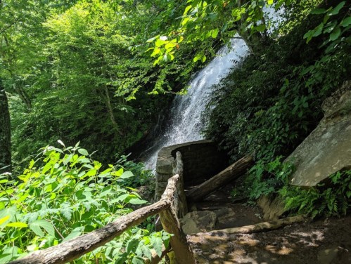 A serene waterfall cascades over rocks, surrounded by lush green foliage and a rustic wooden railing.