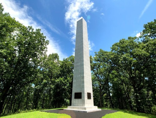A tall, white obelisk monument surrounded by green trees under a blue sky with scattered clouds.