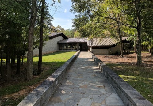 A modern house with a stone pathway, surrounded by trees and greenery, under a clear blue sky.