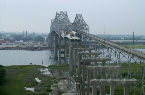 A partially constructed bridge spans a river, with support pillars visible and industrial buildings in the background.