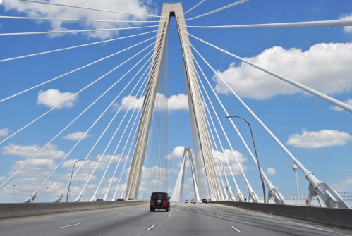 A view of a cable-stayed bridge with a clear blue sky and fluffy clouds, featuring vehicles on the roadway below.