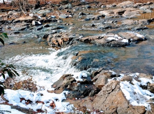 A serene stream flows over rocky terrain, surrounded by patches of snow and bare trees in a tranquil landscape.
